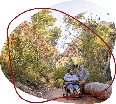 Two men, one in a wheelchair, pauses at the side of a hiking trail.