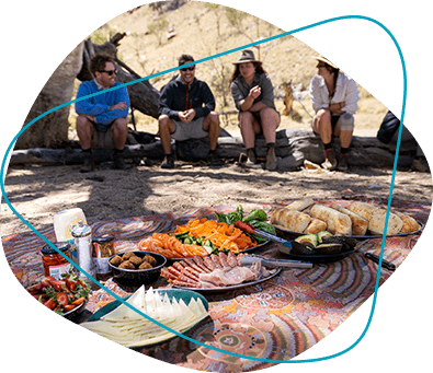 A group of friends sit under a tree appreciating a delicious array of food.