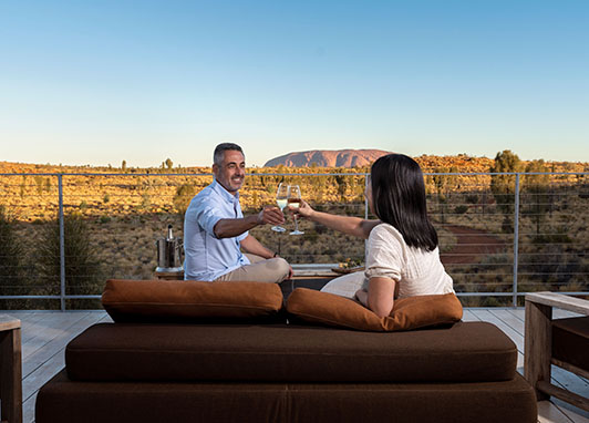 A couple sit on a comfortable veranda toasting glasses of wine with Uluru in the background.