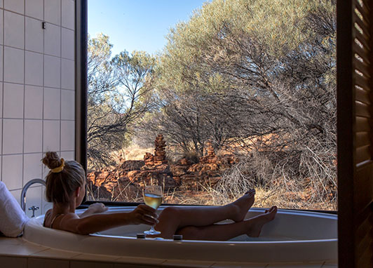 A woman relaxes in a bathtub looking at the desert outside her window while drinking a glass of wine.