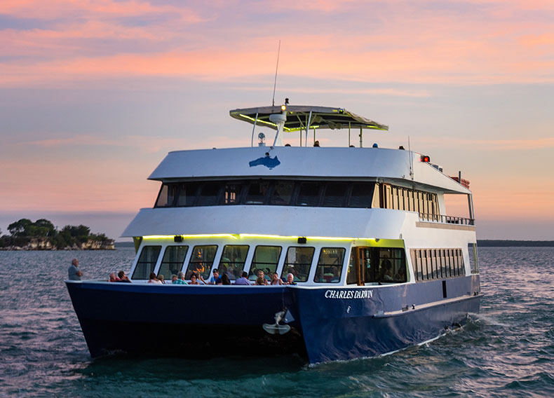A sightseeing ship cruises across a harbour at sunset.