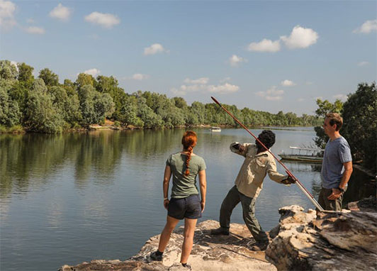 A couple watches an Aboriginal guide demonstrate spear fishing near a river.