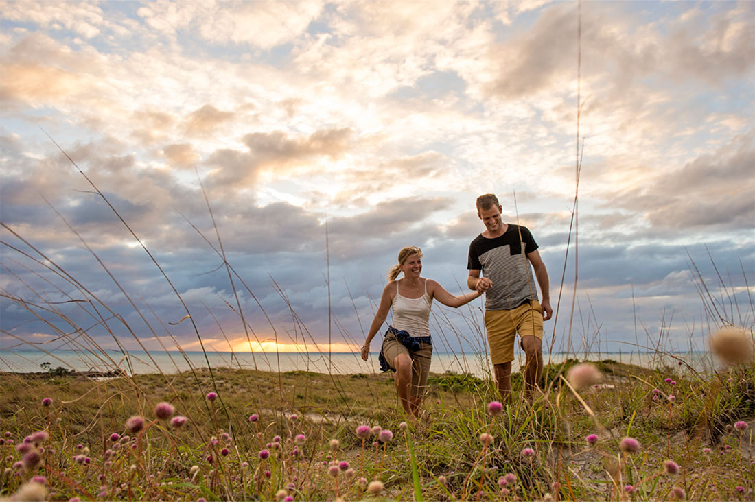 A couple walks through a field of wild grasses and flowers with the ocean in the background.