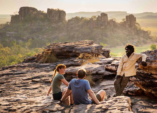 A couple sit on a scenic cliff listening to an Aboriginal guide.