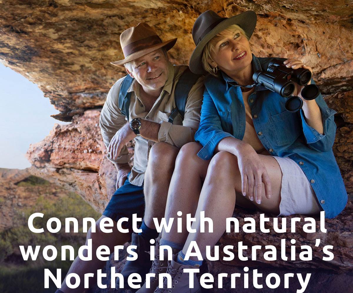 A senior couple sits under a rock outcrop looking at the Outback. A headline reads: Connect With Natural Wonders in Australia’s Northern Territory.