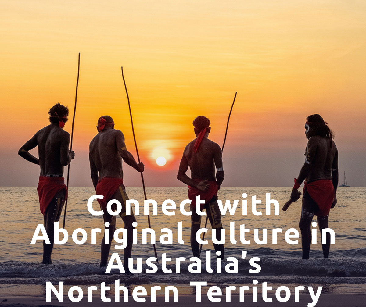 A group of traditionally-dressed Aboriginal men stand on the beach looking at the ocean. A headline reads: Connect With Aboriginal Culture in Australia’s Northern Territory.