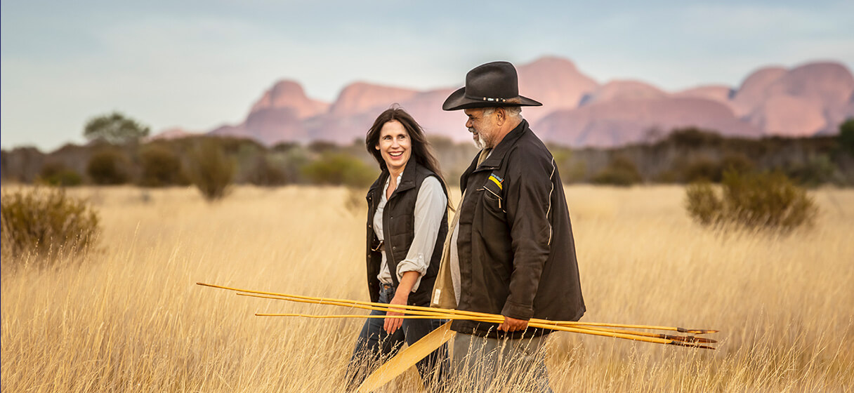 A couple walking through tall grasses carrying aboriginal tools.